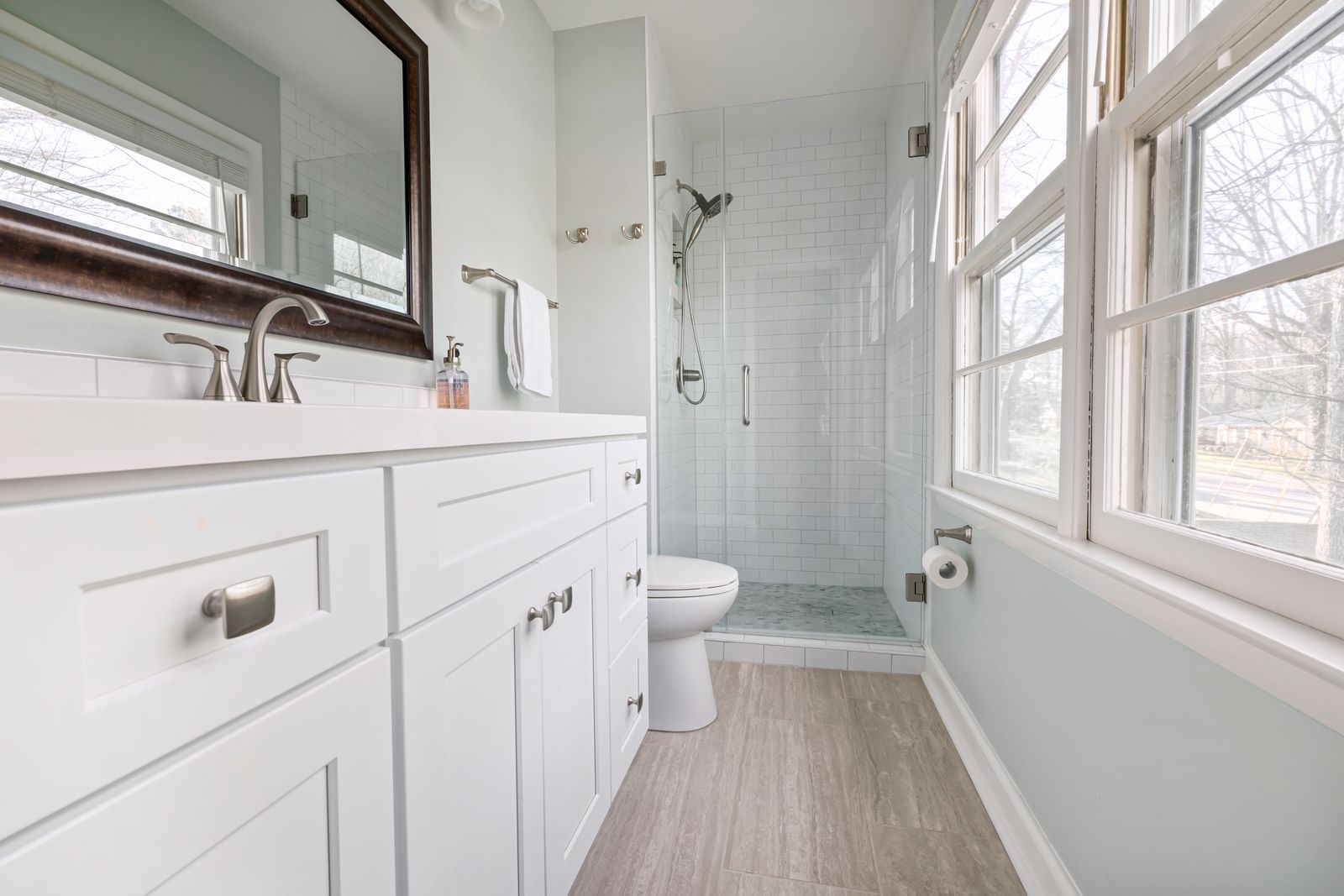 A bright bathroom with a white vanity, a large window, and a walk-in shower