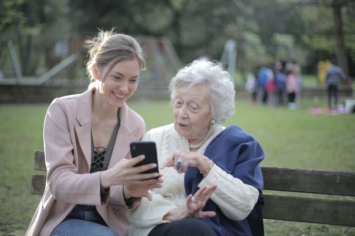 An adult daughter and her elderly mother sitting on a park bench, smiling at a phone together